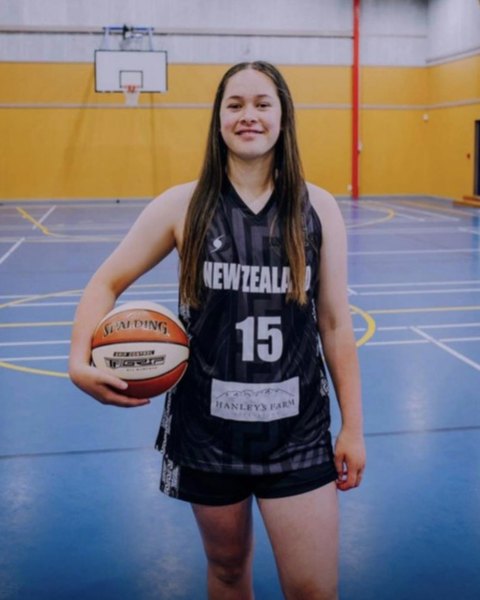 A smiling player in a New Zealand number 15 singlet holds a basketball in a gym.